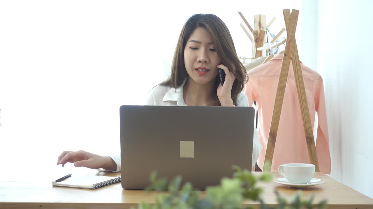 自宅のリビングルームで座ってラップトップで作業している、笑顔の美しい若いアジア人女性。自宅のオフィスで仕事のために電話を使用しているアジア人ビジネスウーマン。自宅での時間を楽しんでいる。
