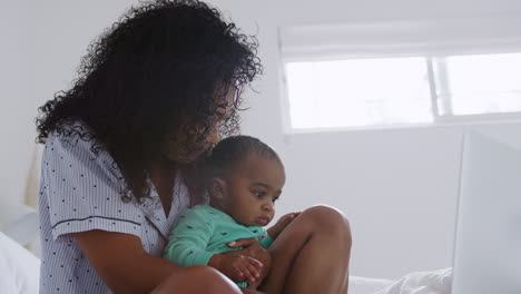 Mother-Wearing-Pyjamas-Cuddling-Baby-Son-In-Bedroom-Whilst-Working-From-Home-On-Laptop