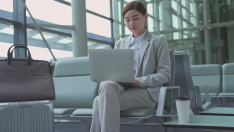 짐을 들고 라운지에 앉아있는 긍정적 인 사업가 여성은 공항에서 비행기의 탑승을 기다리는 동안 태블릿 컴퓨터를 재생합니다. 승객 여행자 여성은 대기실에서 휴대 전화로 소셜 미디어를 확인합니다.