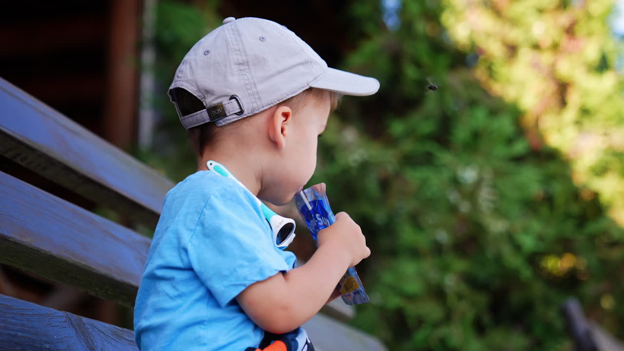 Caucasian boy wearing a cap sits outdoors drinking juice from pack and straw. Low angle view on a lovely baby on the walk.