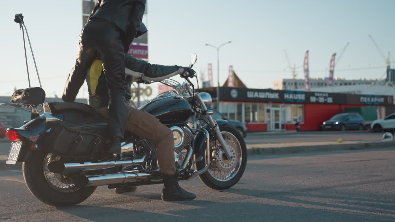 Passenger in leather outfit approaches parked motorcycle preparing to climb behind rider seated on power bike, holding balance as sunlight highlights street and blurred cars in urban background