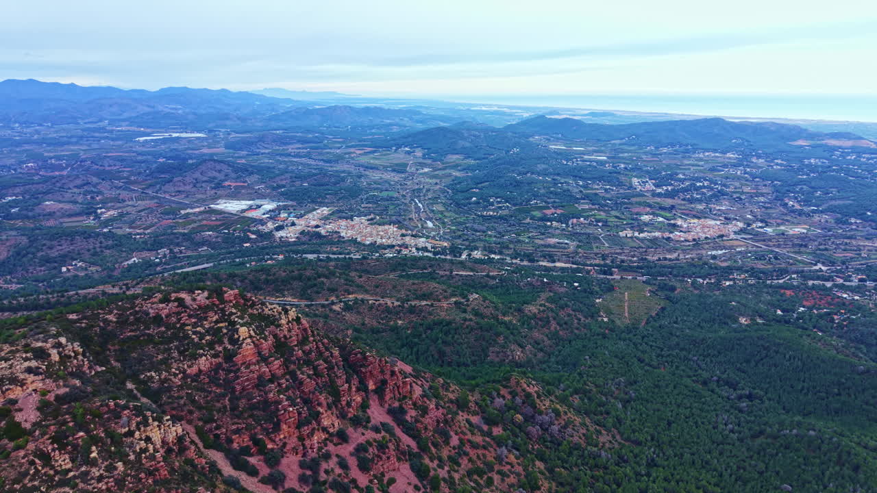 Vista do cume de uma montanha exibindo vales, colinas e uma cidade ao longe em um dia nublado no final da tarde.