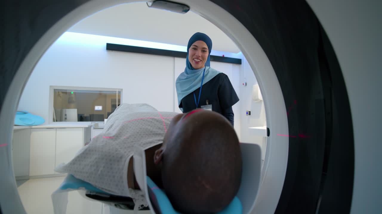 Tecnico di tomografia computerizzata (TC) con hijab che posiziona la testa nel gantry, rassicurando un uomo afroamericano prima della scansione.