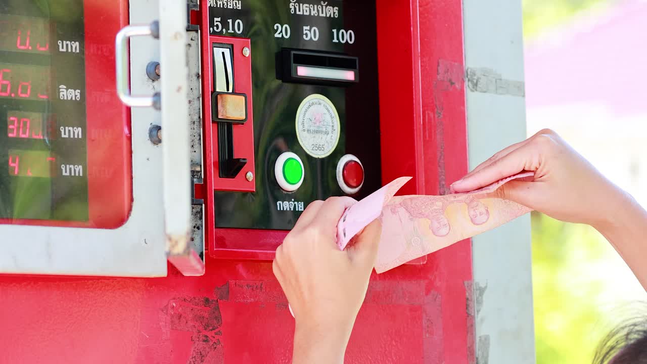 Person inserts cash into red fuel vending machine under bright daylight, close-up perspective, steady camera