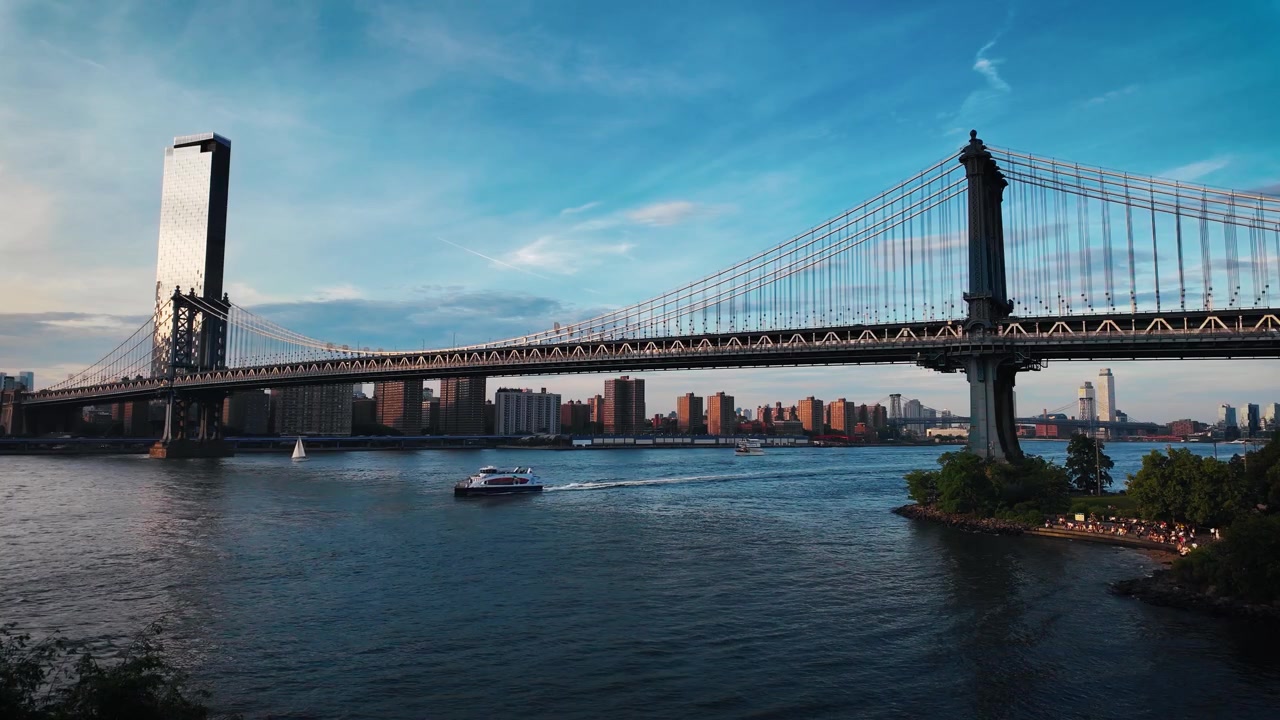 Vista panoramica statica del Ponte di Manhattan sopra l'East River con Roosevelt Island, barche di passaggio e nuvole colorate al tramonto che incorniciano lo skyline urbano di New York