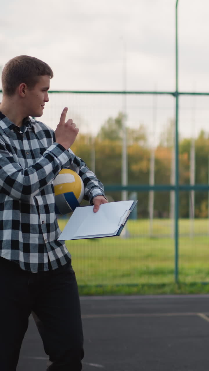 entrenador de voleibol sosteniendo la pelota bajo el brazo y el libro de récords en la mano, instruyendo a las principiantes que lo están mirando atentamente con sonrisas cálidas durante una sesión de entrenamiento al aire libre