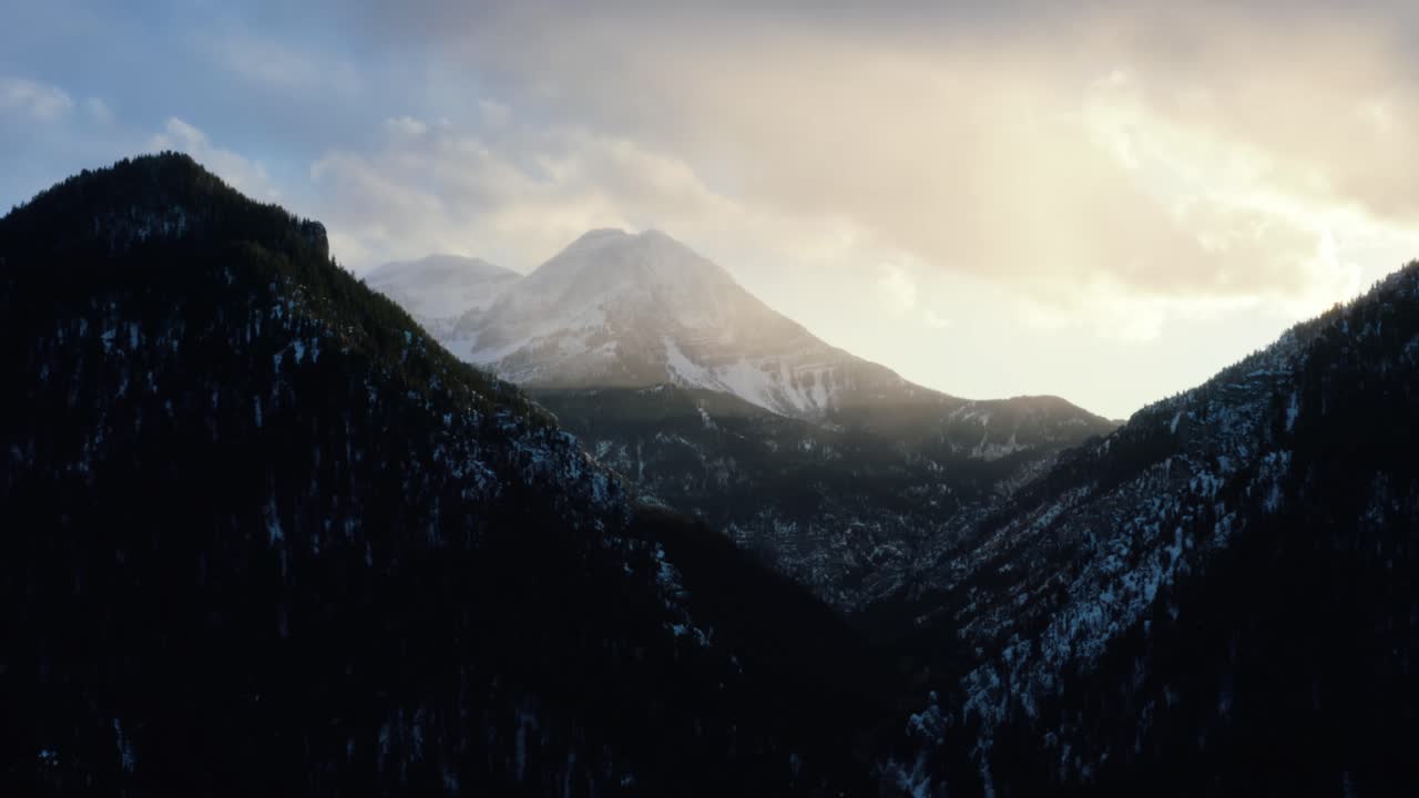 Static aerial drone shot of a winter landscape of Mount Timpanogos in the background surrounded by a pine tree forest during sunset from the frozen Tibble Fork Reservoir in American Fork Canyon, Utah