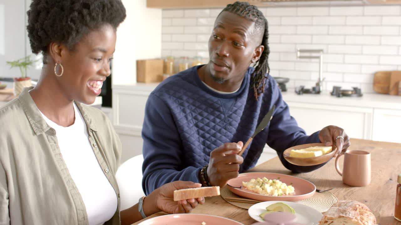 Smiling African american couple enjoying breakfast together, serving food at home kitchen table