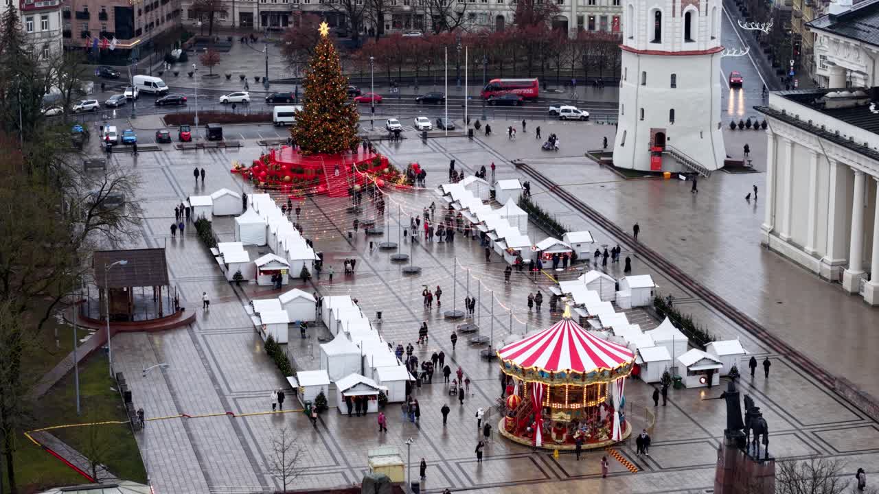 Aerial daytime view of the Christmas market in Vilnius, showing the decorated tree, carousel, vendor cabins, and Cathedral Square activity on a rainy festive afternoon