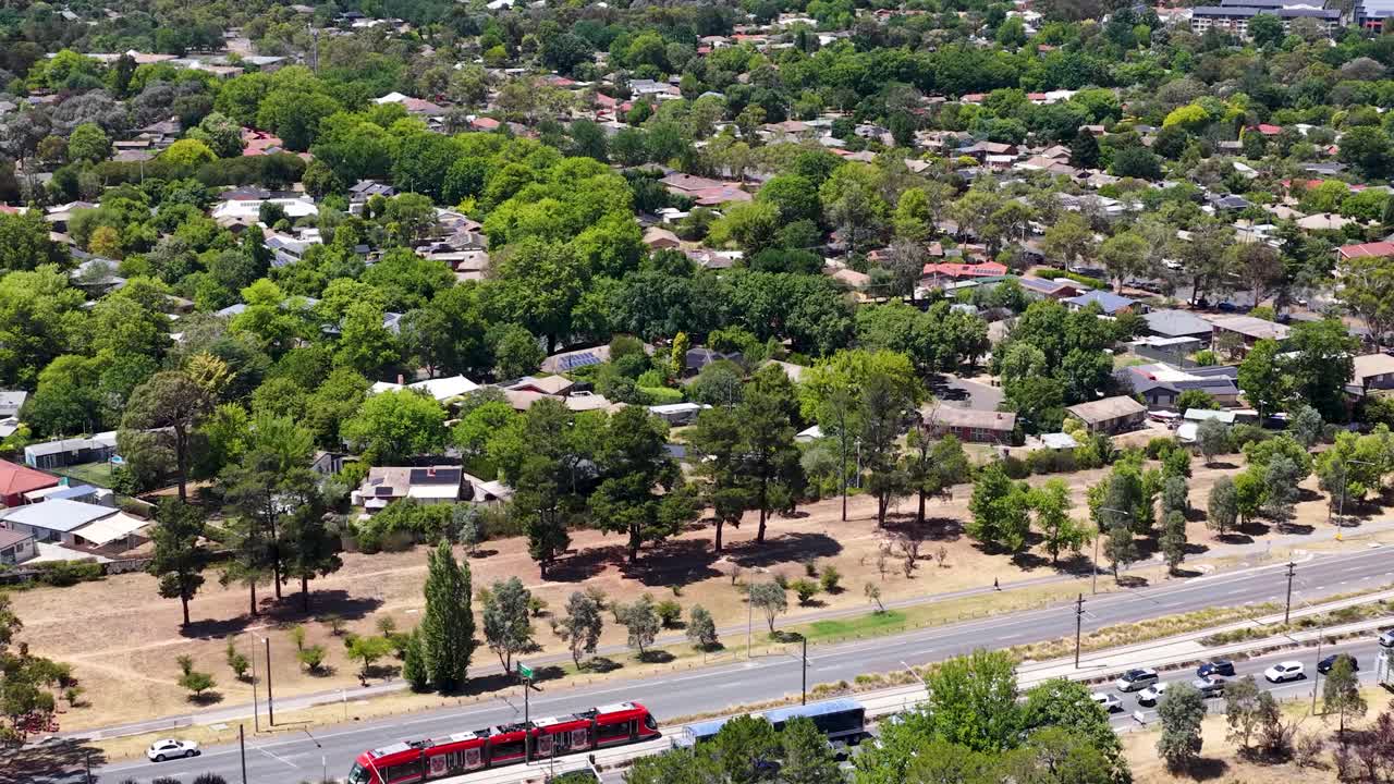 Ripresa aerea dall'alto che segue un tram rosso che si muove attraverso un sobborgo residenziale verde a Canberra