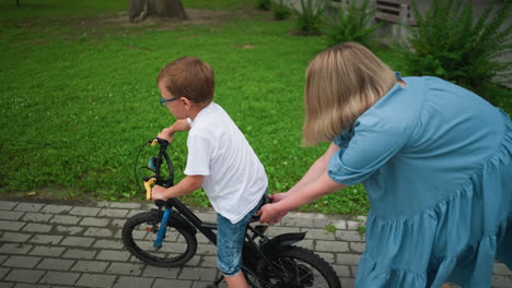 a woman holds the seat of her child s bicycle, helping him ride along a paved walkway next to lush greenery