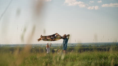 a woman in a blue dress holds a plaid scarf as it dances in the wind on a grassy field. two children in white shirts play nearby, one bending down and the other reaching out to touch the flowing scarf