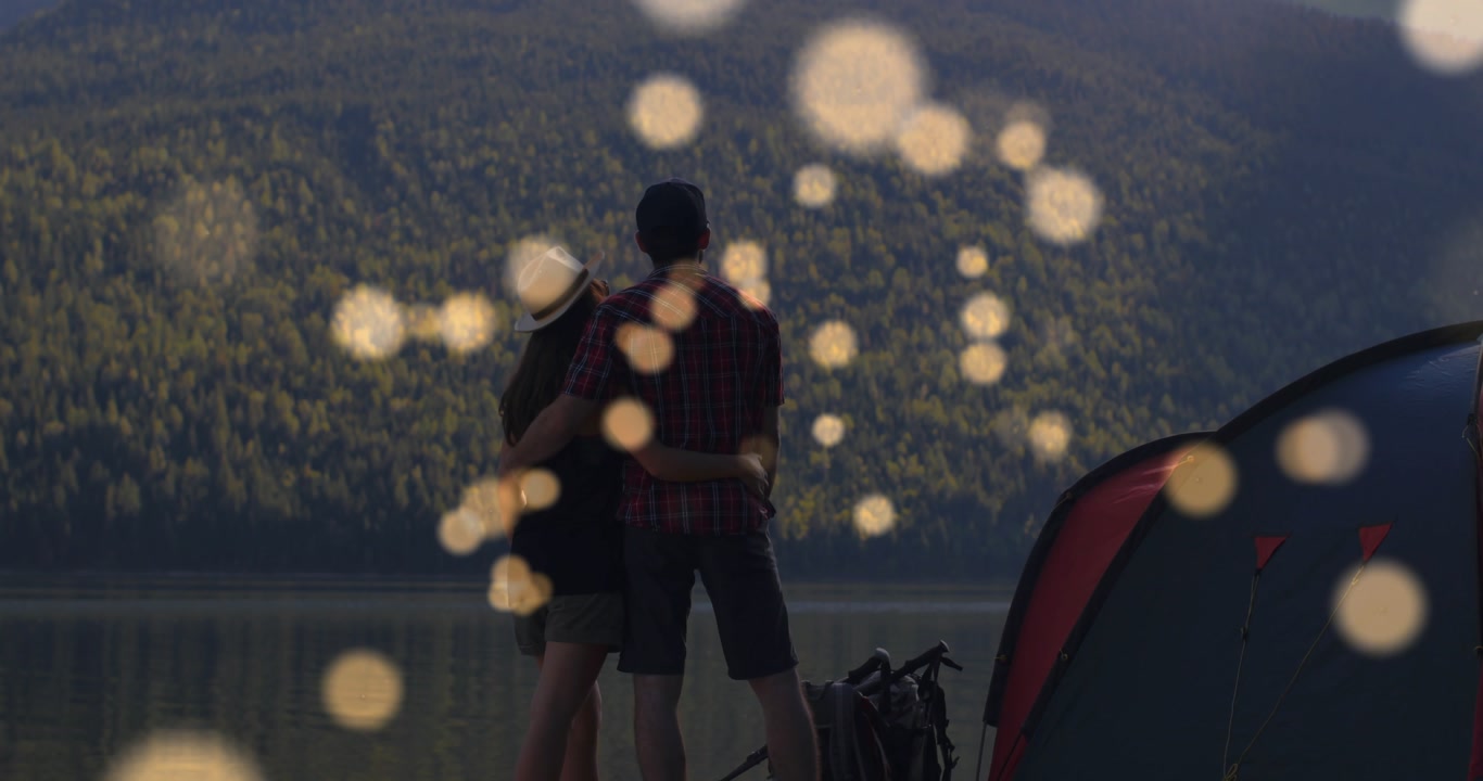 Campers standing shore watching lake, bokeh drifting over them while leaning gesturing, tent bikes