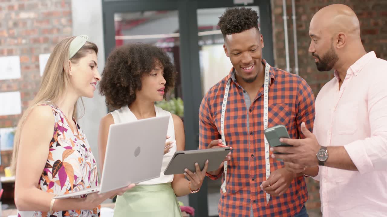 diseñadores diversos felices con tabletas, teléfonos inteligentes y computadoras portátiles hablando en un estudio de moda, en cámara lenta