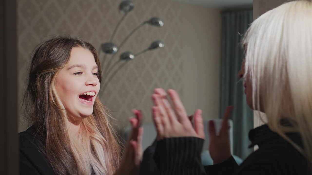 close up of two women with casual attire smiling as they clap palms in friendly high-five gesture with blurred room decor and lamp in background