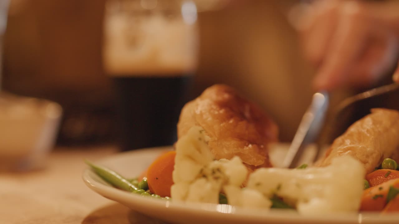 A person uses a knife and fork to cut into a steak pie served with mixed vegetables in a warmly lit pub setting, with a pint of dark beer visible