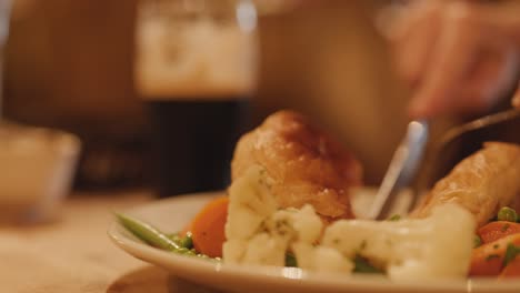 a person uses a knife and fork to cut into a steak pie served with mixed vegetables in a warmly lit pub setting, with a pint of dark beer visible