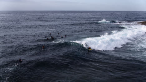 Seguimiento-Aéreo-De-Un-Grupo-De-Surfistas-Y-Bodyboarders-Montando-Olas-Cerca-De-Las-Rocas