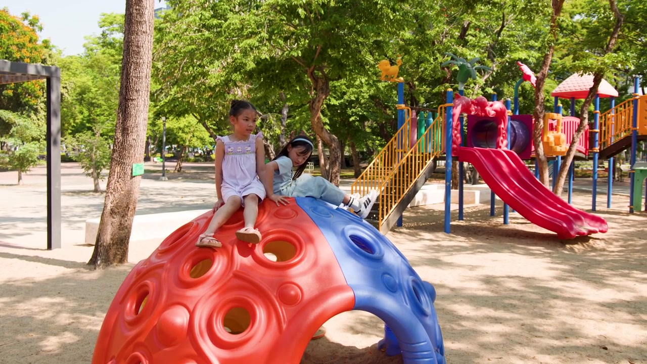 Dos niñas asiáticas se sientan en una colorida cúpula de escalada en un soleado parque infantil de Lumpini Park.