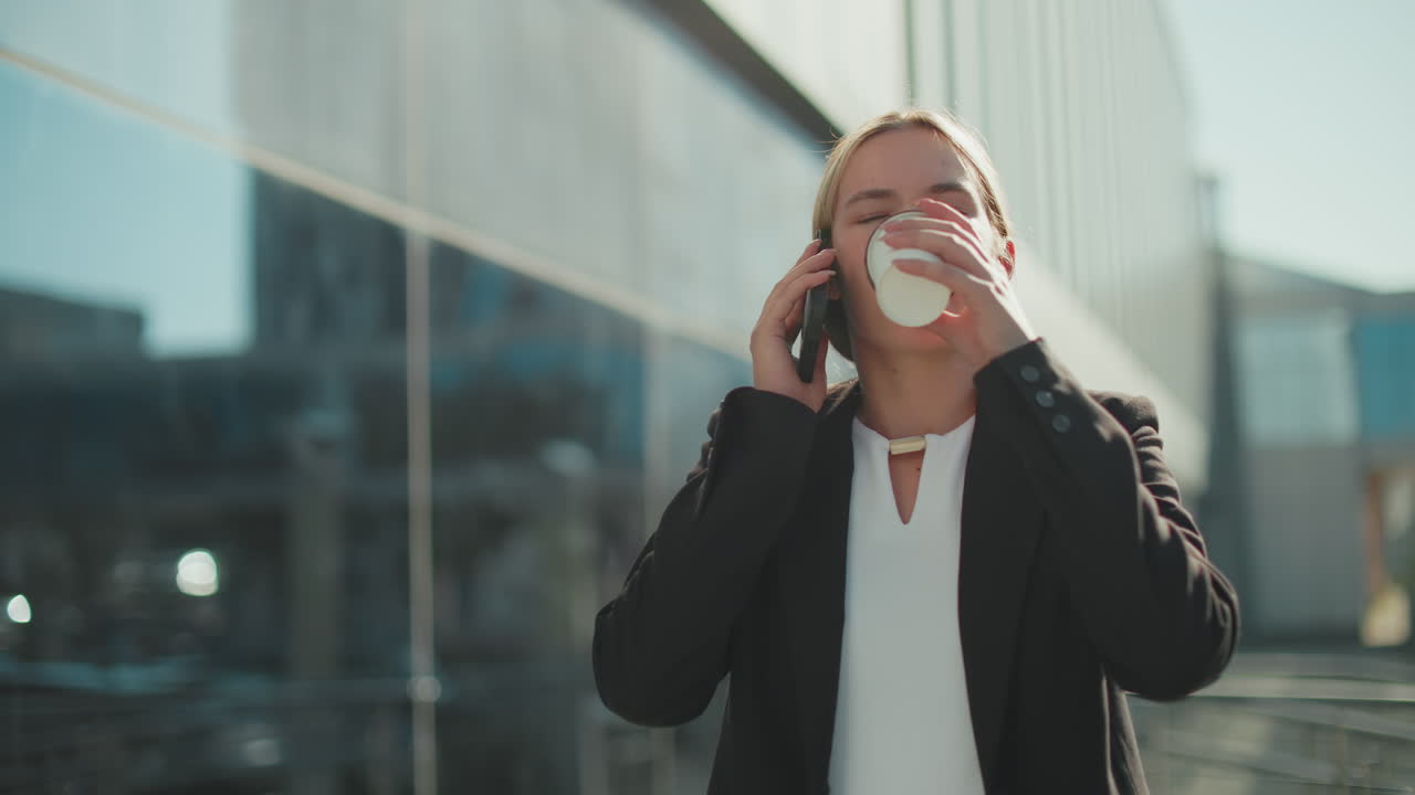 Banker on break taking juice while on phone call, walking past glass facade building under bright daylight, reflecting surrounding cityscape with soft sunlight illuminating her