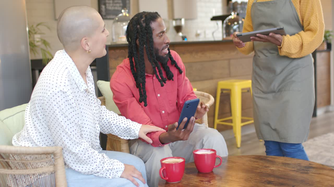 Diverse couple scrolling phone and laughing at message before barista showing tablet at coffee shop