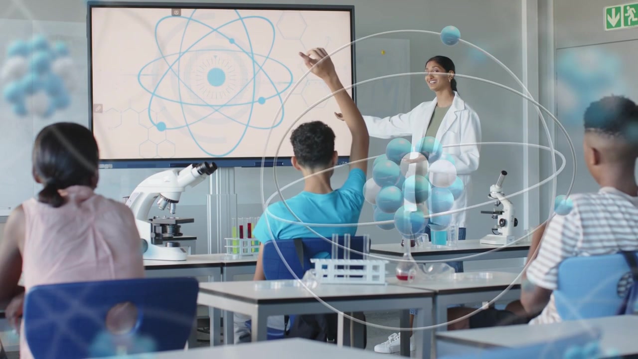 Lærer peker på skjerm i realfagsrom, 3D-atom vises, student rekker opp hånden for å avklare.