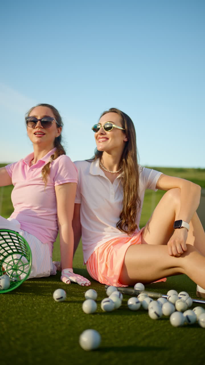 Two women standing on the golf course near a bucket with spilled balls. Vertical