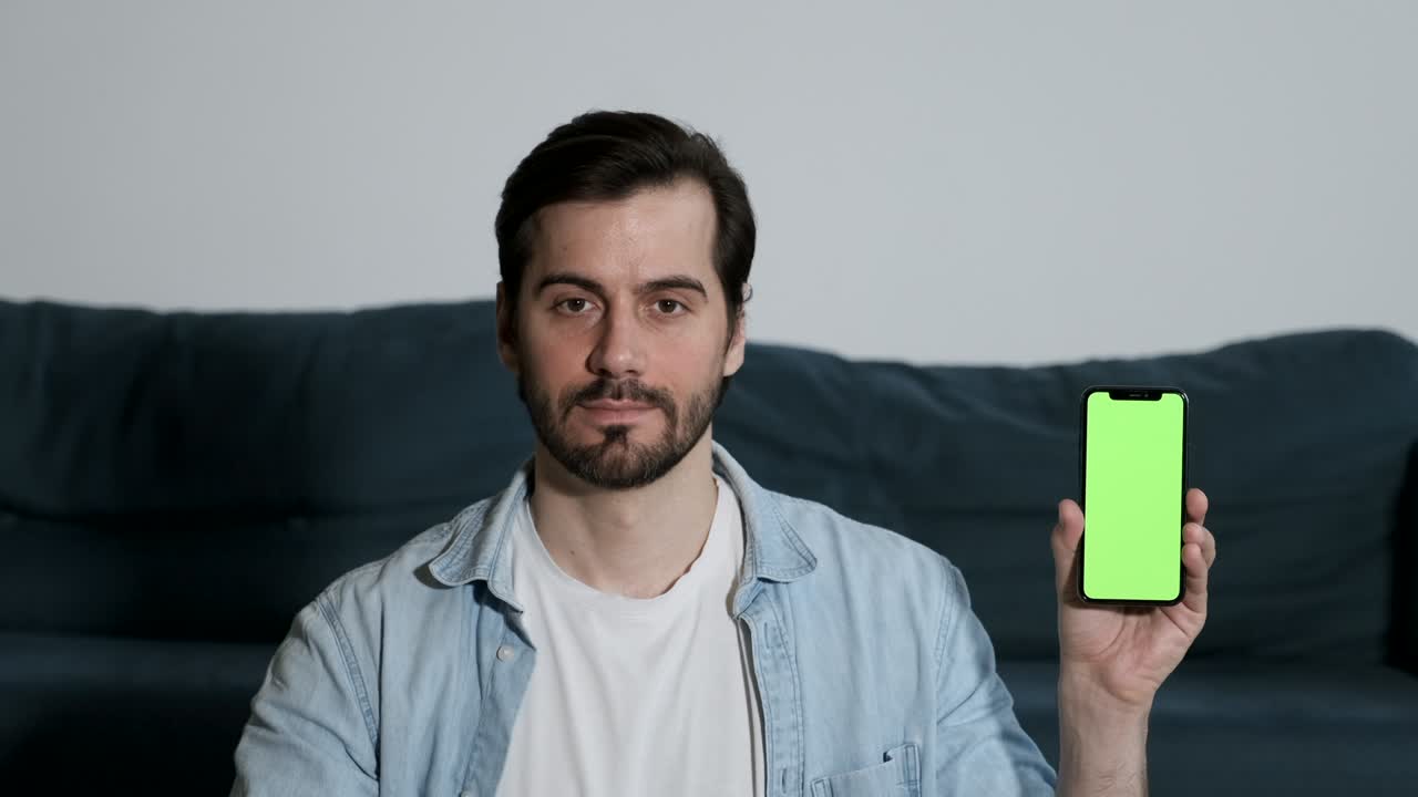 젊은 남자가 카메라를 보고 새로운 응용 프로그램의 녹색 화면 통합으로 스마트폰을 보여줍니다.