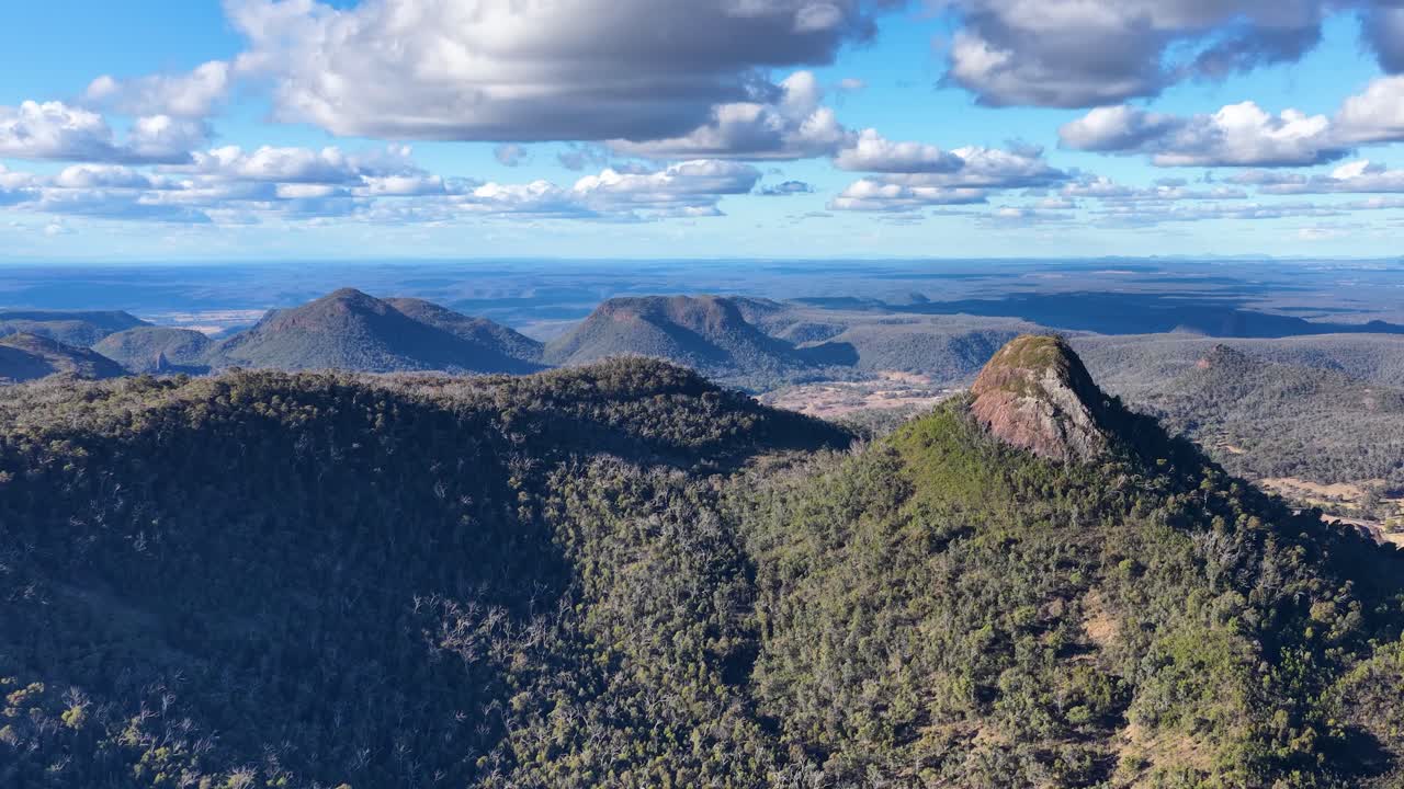 Drone footage smoothly pans across rugged Warrumbungle mountain peaks under partly cloudy skies, highlighting forested slopes and dramatic afternoon lighting in rural Australia