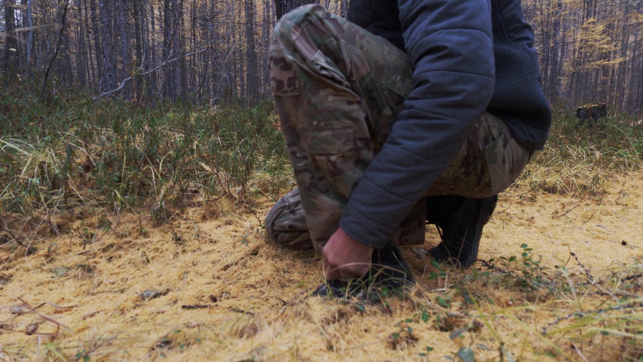 Una persona con pantaloni mimetici e una giacca scura si accovaccia in una radura della foresta per allacciarsi gli scarponi neri, poi si alza ed esce verso sinistra attraversando erba secca giallastra e aghi di pino