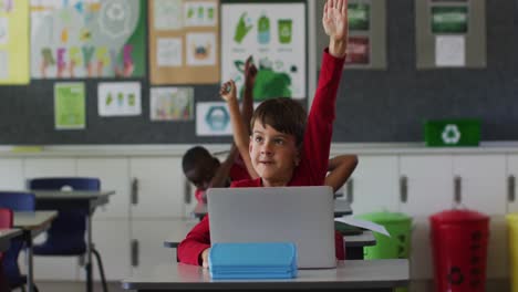 retrato de un escolar caucásico feliz con una computadora portátil sentado levantando la mano para responder preguntas