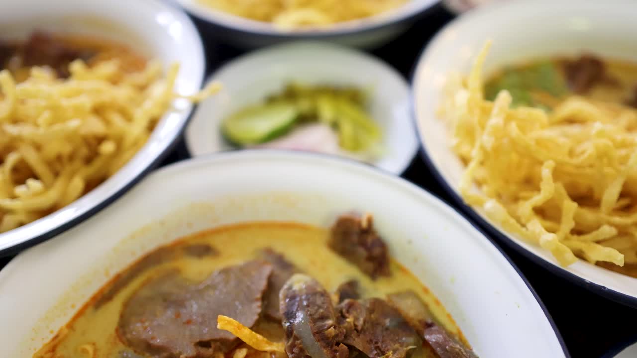 Multiple bowls of beef curry noodles with crispy toppings and side garnishes are shown in a brightly lit, close-up sequence, highlighting vibrant textures and colors