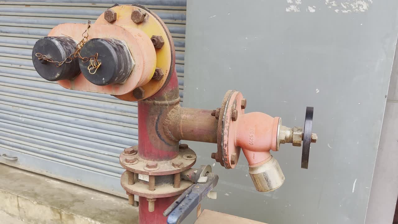 Camera circles around a red outdoor fire hydrant system with control valves, bolts, and fittings near a shuttered building wall