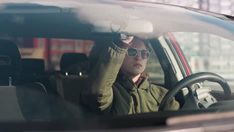 young man wearing winter jacket in parked car lifts sun visor upward while seated in driver seat on bright day with reflections on windshield and urban buildings visible in background