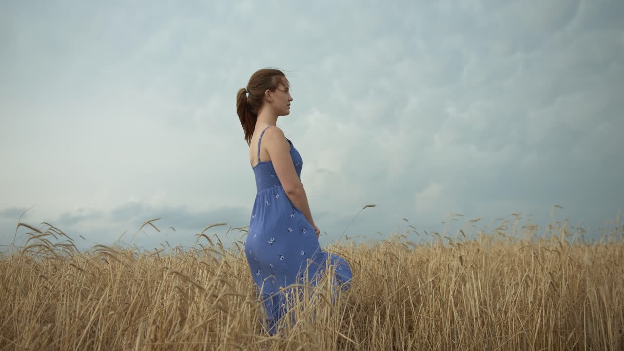 A young woman reflecting in nature, wearing a blue dress standing in a wheat field under moody, storm clouds in summer.