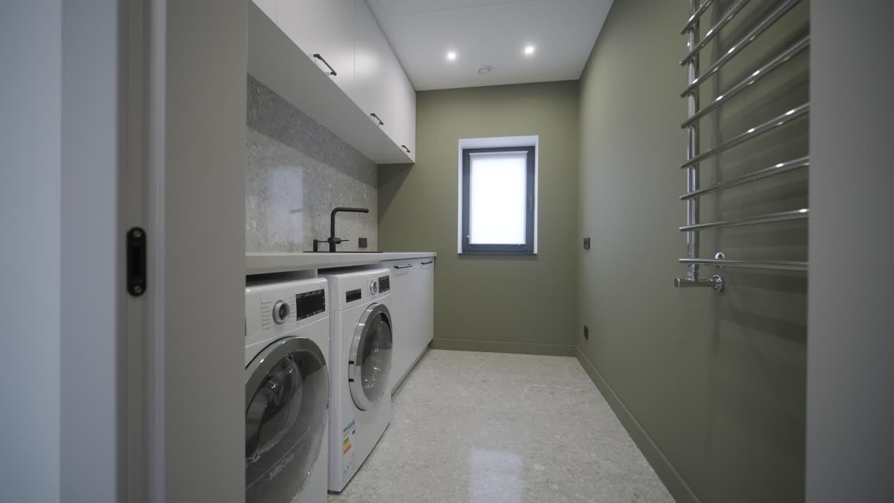 una lavandería funcional equipada con electrodomésticos modernos, una encimera de granito y un pasillo de toallas calentado, colocado contra una pared de color verde olivo y un piso de terrazo