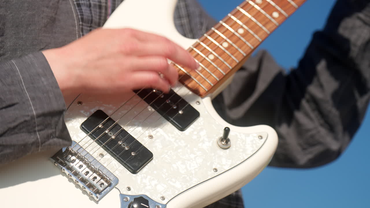 Mani che suonano una chitarra elettrica bianca all'aperto sotto un cielo blu, con focus sulla pennata, sul dettaglio della tastiera e sull'energia della performance musicale dal vivo, primo piano