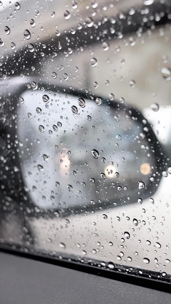 Gocce di pioggia sul finestrino dell'auto, specchietto laterale sfocato, traffico cittadino, luce diurna, messa a fuoco selettiva, leggero movimento della telecamera