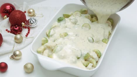 pouring creamy bechamel sauce over brussels sprouts and cauliflower with christmas balls on the side