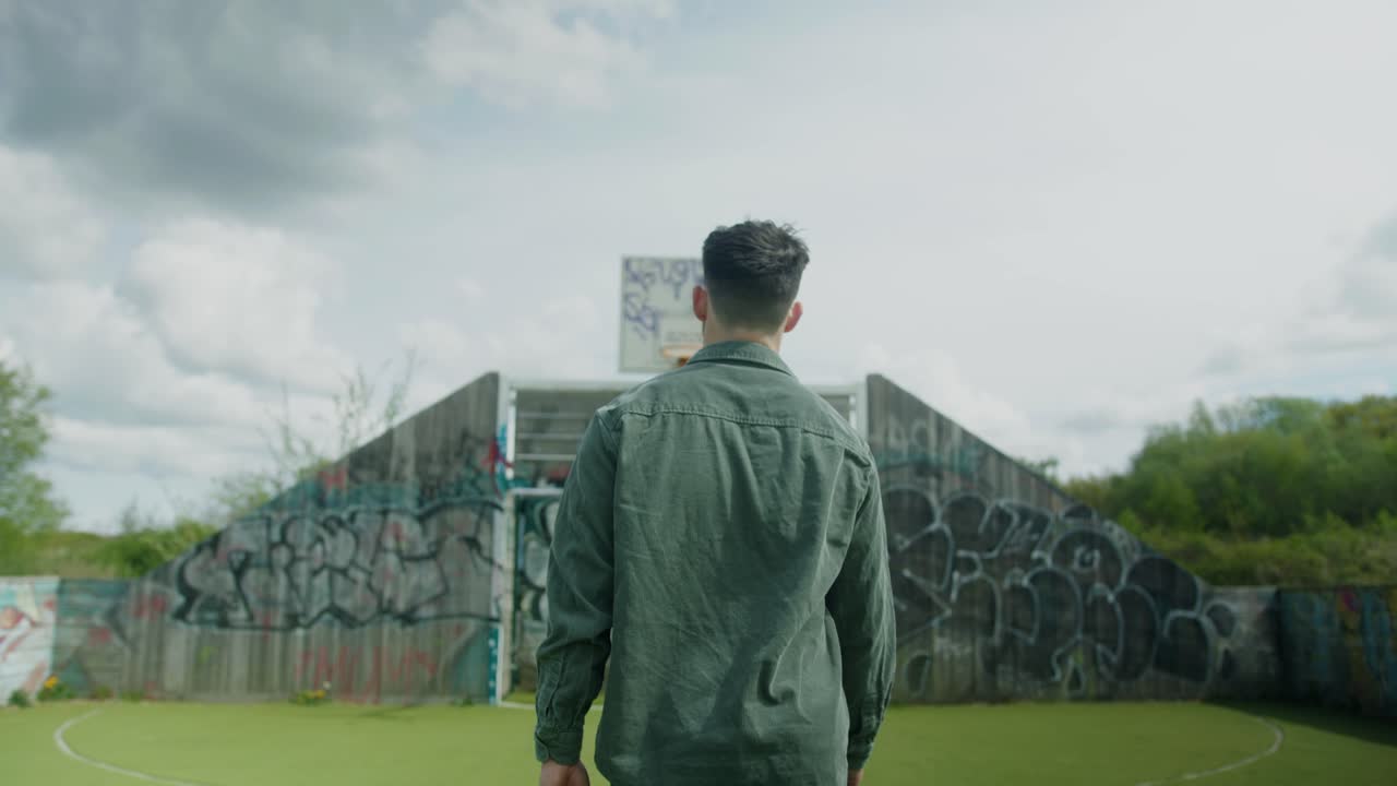 Tracking rearview of man walking on basketball court, overcast sky, framed by greenery and hoop