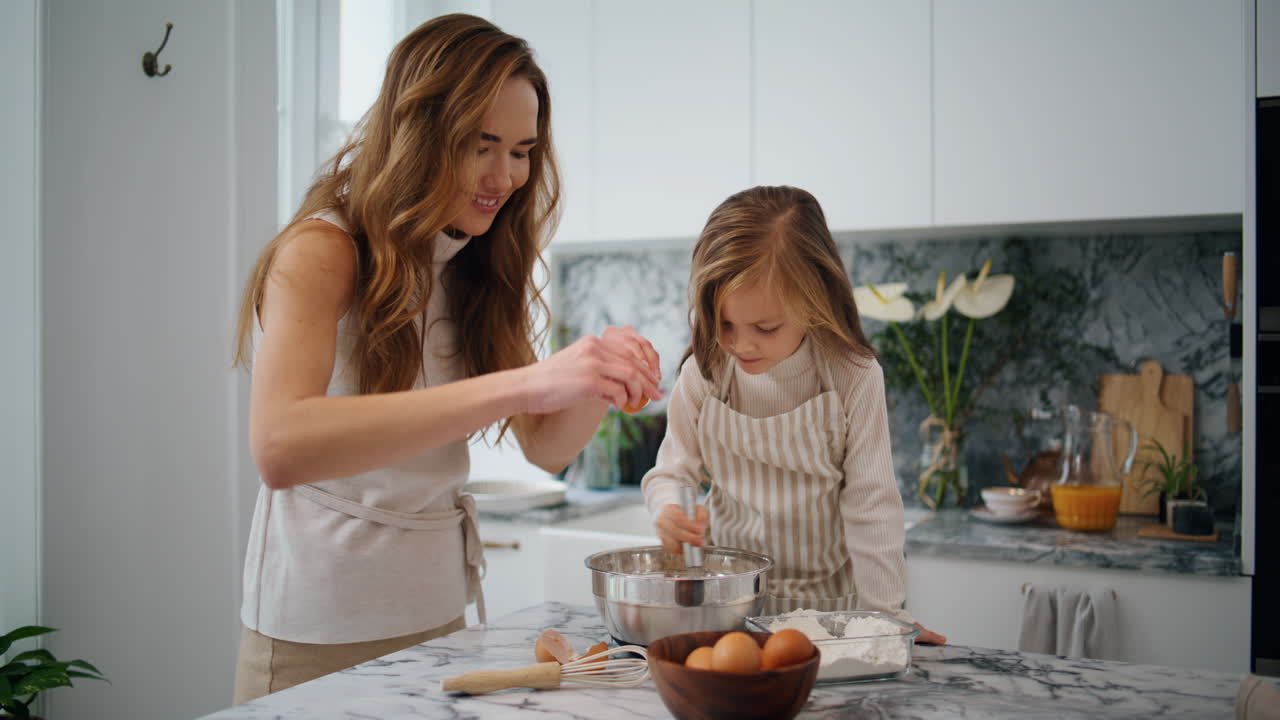 Smiling mother daughter breaks eggs at kitchen. Child mixing ingredients at bowl