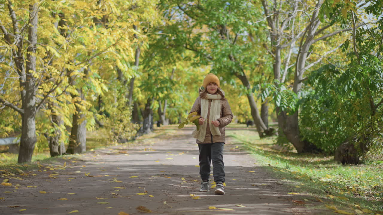 side view of young kid strolling down leaf strewn path holding yellow autumn leaf while wearing grey joggers, chunky sneakers, mustard beanie and knitted scarf under golden tree canopy