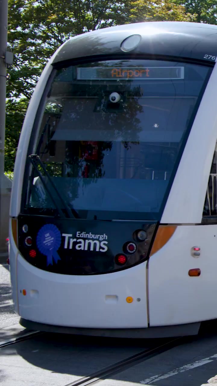 Tram che viaggia lungo i binari della città, passando davanti a negozi e panchine, sotto una luce diurna brillante con telecamera stabile.