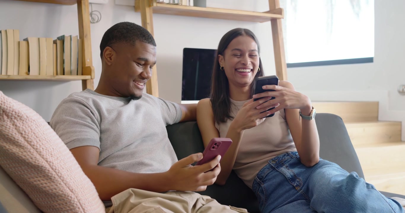 Casal afro-americano sentado no sofá, a mulher levantando o telefone mostrando uma piada, o homem rindo.
