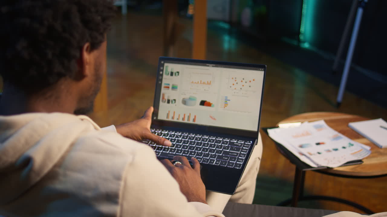 freelancer afroamericano que trabaja desde casa usando gráficos financieros, revisando datos estadísticos para comparar números de oferta y demanda. usando gráficos de negocios y escribiendo en el bloc de notas. cámara b.