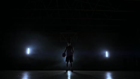 The-basketball-player-stands-on-a-dark-Playground-and-holds-the-ball-in-his-hands-and-looks-into-the-camera-in-the-dark-with-a-backlit-in-slow-motion-and-around-smoke