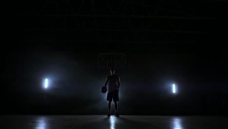 the basketball player stands on a dark playground and holds the ball in his hands and looks into the camera in the dark with a backlit in slow motion and around smoke