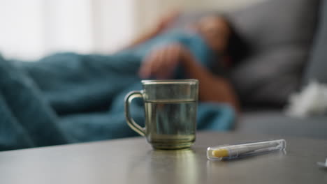 water inside glass cup on table with thermometer and blurred background showing restless person on couch, dim light indicating a sick day, recovery, and comfort in peaceful setting