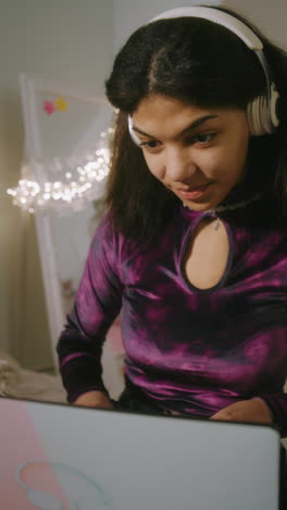 Vertical-view-of-teen-girl-in-headphones-typing-on-keyboard-and-chatting-with-friend-online-on-laptop-sitting-in-her-cozy-bedroom.-African-American-girl-spending-leisure-time-and-having-fun-at-home.