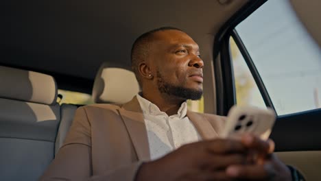 confident man businessman with black skin color in a brown suit drives through the city looking out the window and chatting on his white phone while waiting for arrival